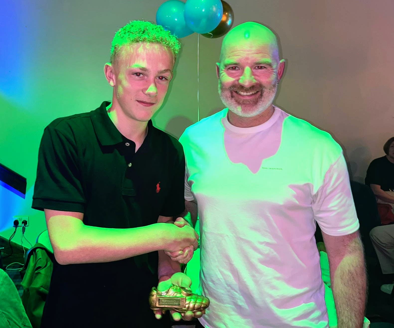 A young award recipient holding a golden boot trophy beside a coach