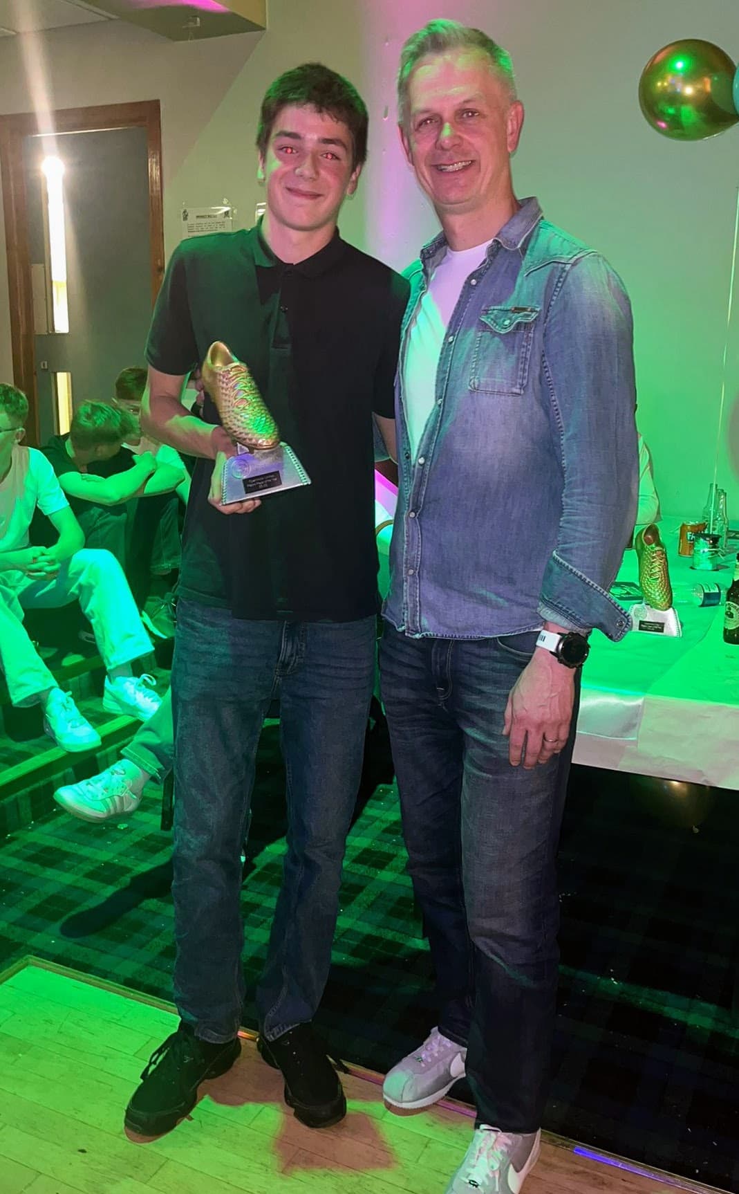 A young award recipient holding a golden boot trophy beside a coach